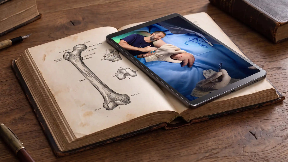 Tablet reproduzindo vídeo de treinamento cirúrgico apoiado sobre um livro de anatomia antigo aberto.